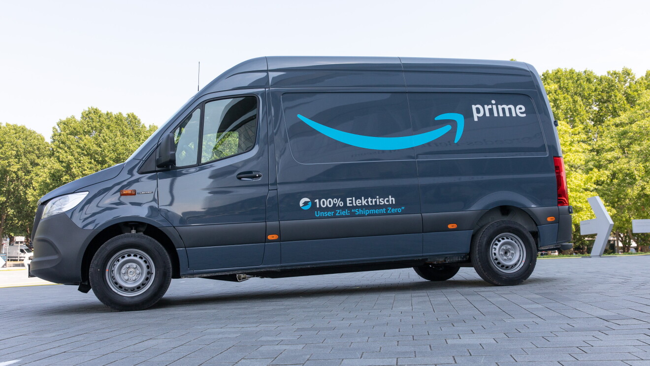 Una furgoneta eléctrica de Mercedes-Benz con el logo Prime de amazon y debajo en alemán 100% eléctrica. La furgoneta sin conductor está en una plaza con árboles en el fondo. 