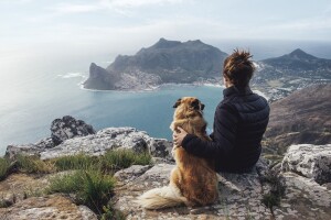 A dog and female owner sitting on a ledge, overlooking an ocean with rock and peninsula outcropping.