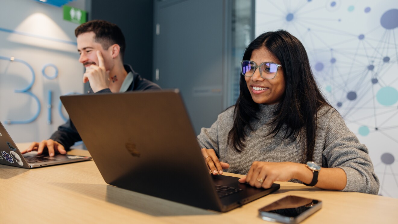 An man and woman sitting at a table in a modern collaborative workspace working on their laptops side by side, smiling