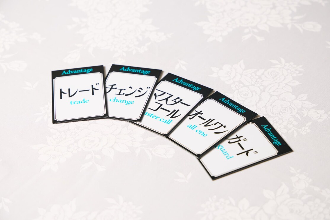 バラ模様が入った白いテーブルクロスの上に黒い縁取りのカードが5枚広げられている。カードに書かれた文字は左からトレード、チェンジ、マスターコール、オールワン、ガード