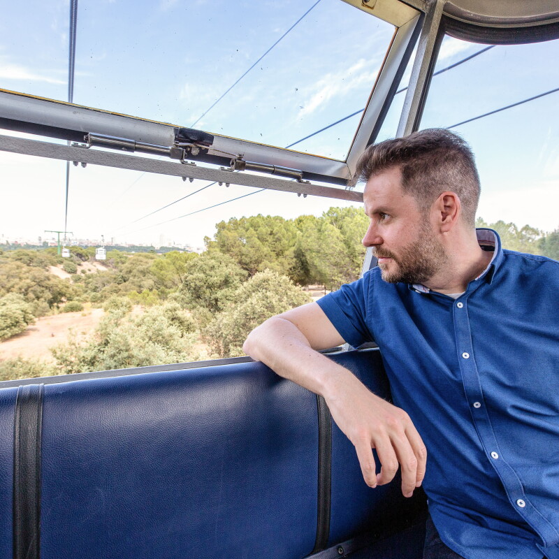 En el teleférico de Madrid, Paul Pen mirando al horizonte. La ventana de la cabina está abierta y  Paul tiene un brazo reposando en la silla. El escritor lleva una camisa azul y el pelo corto. 