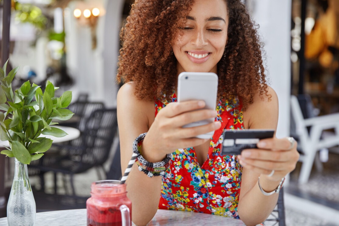 Foto de uma mulher, sorrindo, segurando um celular e um cartão de crédito faz compras online
