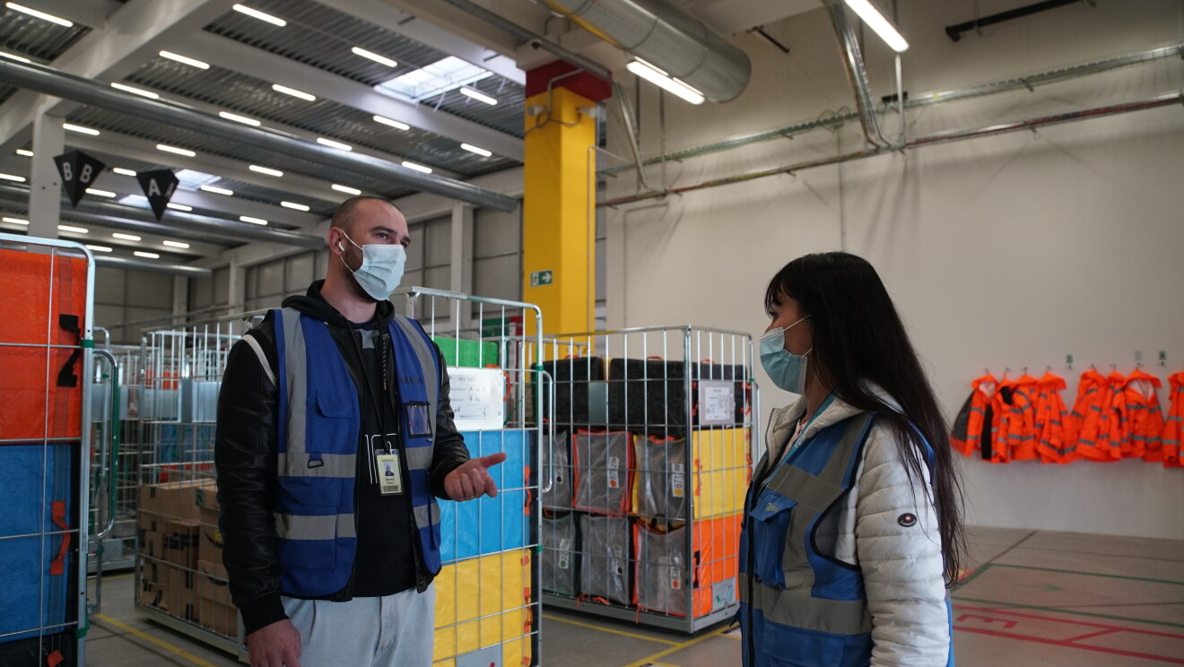 Due dipendenti di Amazon, un uomo e una donna, parlano tra loro in un magazzino. Alle loro spalle, dei carrelli con pacchi di Amazon e dei giubbini di sicurezza rossi appesi al muro. 
