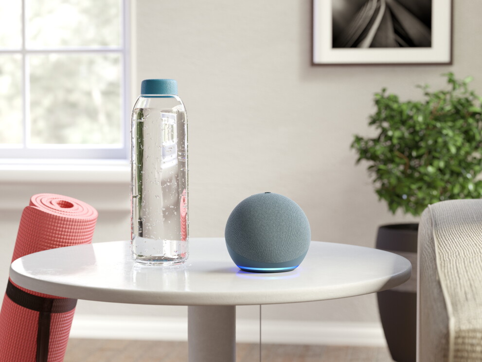 An Amazon Echo dot on a small table next to a bottle of water, leaning on the table is a pink yoga mat.