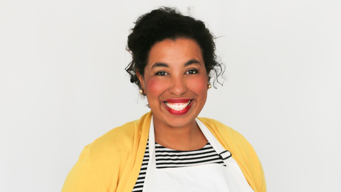 An image of a woman smiling for a photo against a white background. She is wearing a yellow shirt and a white apron.