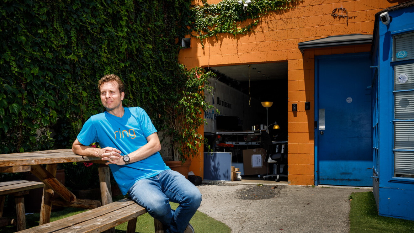 Jamie Siminoff, founder of Ring, sits at a picnic table at Ring headquarters in Santa Monica, CA. He is in a blue t-shirt and jeans, and is looking to the left. 