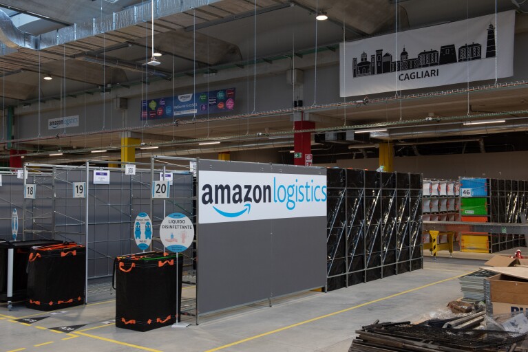 Foto di un'area di un magazzino Amazon. Su un pannello al centro si vede il logo di Amazon Logistics, mentre in fondo uno striscione con il profilo di una città e la scritta Cagliari