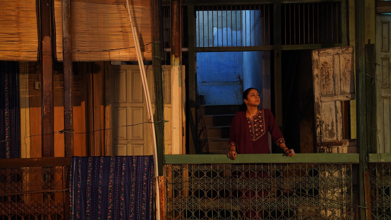 Person in embroidered burgundy tunic on residential balcony during golden hour, framed by traditional wooden architecture
