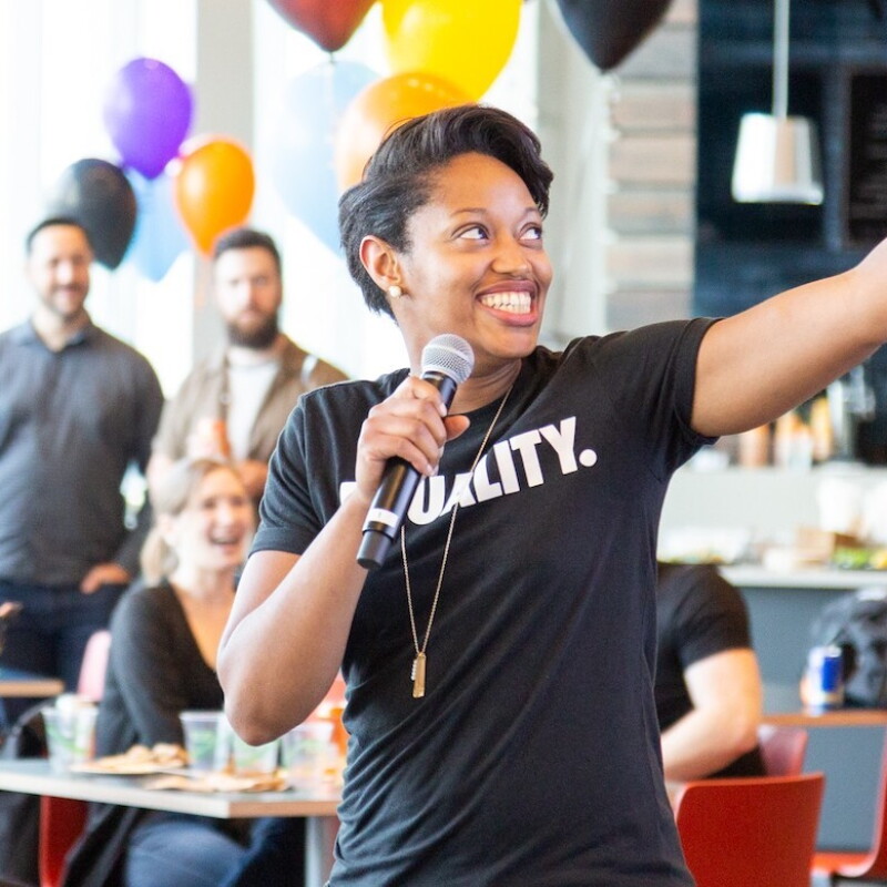 A Black woman wearing a shirt that says "Equality" speaks into a microphone while gesturing off camera. She's speaking to a brightly decorated room full of people.