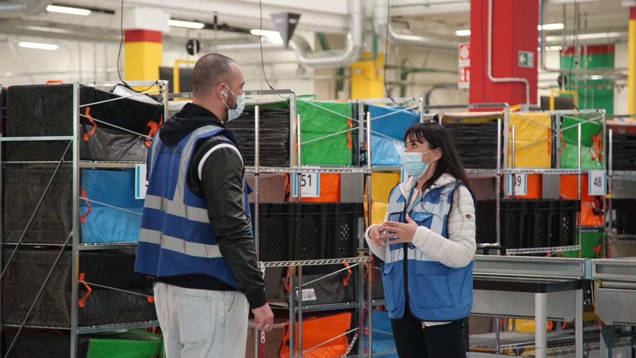 Due dipendenti di Amazon, un uomo e una donna, parlano tra loro in un magazzino. Alle loro spalle, dei carrelli con pacchi di Amazon. 