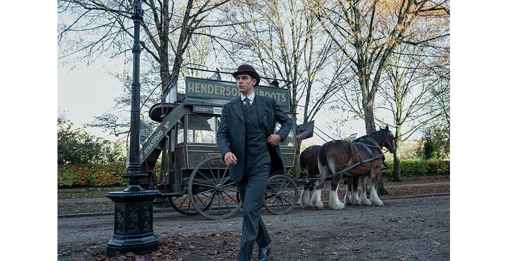 街路樹と街灯が見える屋外。三つ揃いのスーツを着てさっそうと歩く男性。帽子をかぶっている。背後に二頭立ての馬車がとまっている。