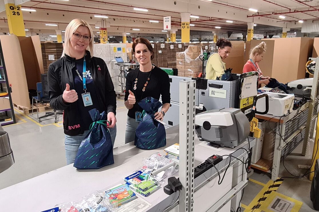 Zwei Mitarbeiterinnen von Amazon mit blauen Taschen in einem Logistikzentrum für eine Spenden-Aktion.