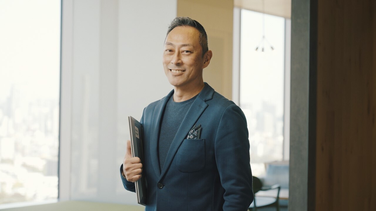 オフィスの窓辺に立つジャケットを着た男性。右手にPCを抱えている