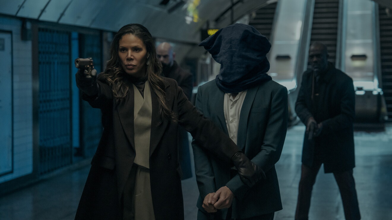 Citadel S2: Woman in brown coat pointing gun while escorting hooded figure in subway station