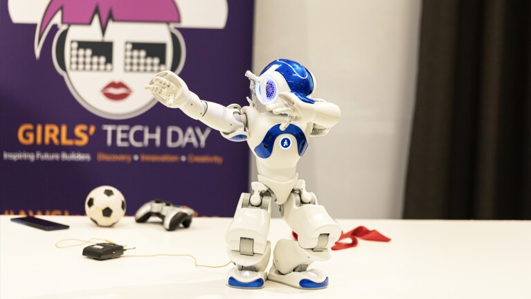 A robot sitting on a table with a soccer ball and remote contol behind it. A poster reads, "Girls' Tech Day" behind the table.