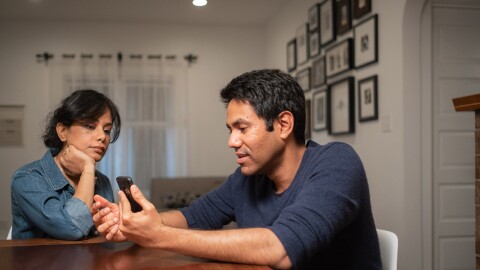 An image of a man and a woman sitting at their kitchen table. The man is video calling someone on his phone while the woman listens in the background.