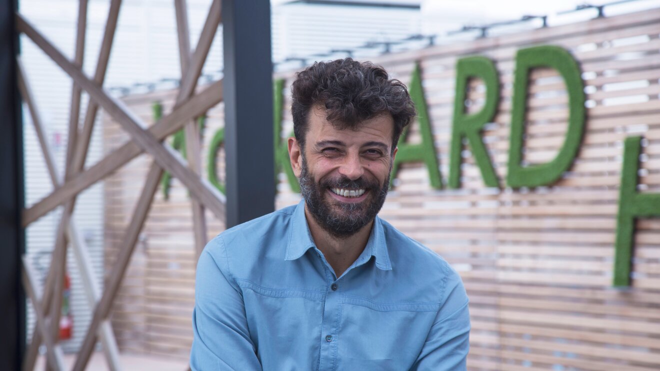 Alessandro Saccon, PR Manager di Amazon, sorride in primo sul terrazzo degli uffici Amazon di Milano. Sullo sfondo uno scorcio del terrazzo, con alcune lettere del motto 'Work hard' di Amazon.