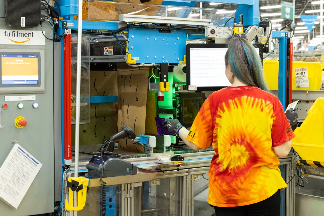 Eine Frau mit gelben und roten T-Shirt steht vor der Verpackungsmaschine in einem Amazon Logistikzentrum.