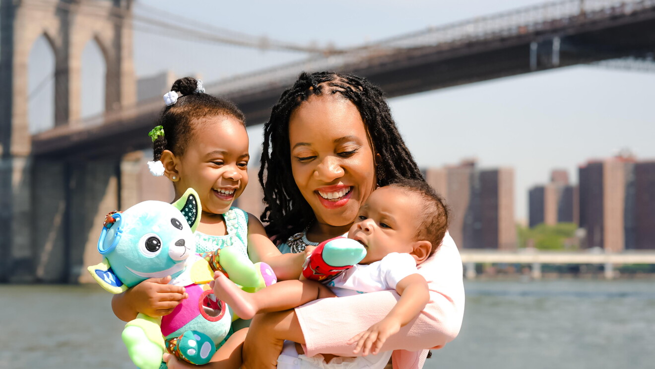 A woman holds a baby and a young child in her arms.  The group is standing in front of a river and a bridge
