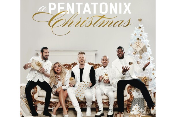 The members of Pentatonix sit on an oppulent ivory sofa with gold trim. A male in white button down with jacket and black pants sits next to a blonde woman leaning forward. She wears a white dress and flats. A man holds a gift, and wears white pants, black tee and white vest. A man in white tuxedo sits next to him and holds a gift. A man throws a gift up in the air. He wars a white tuxedo jacket with black and gold pocket square, white button down and tie and black pants.