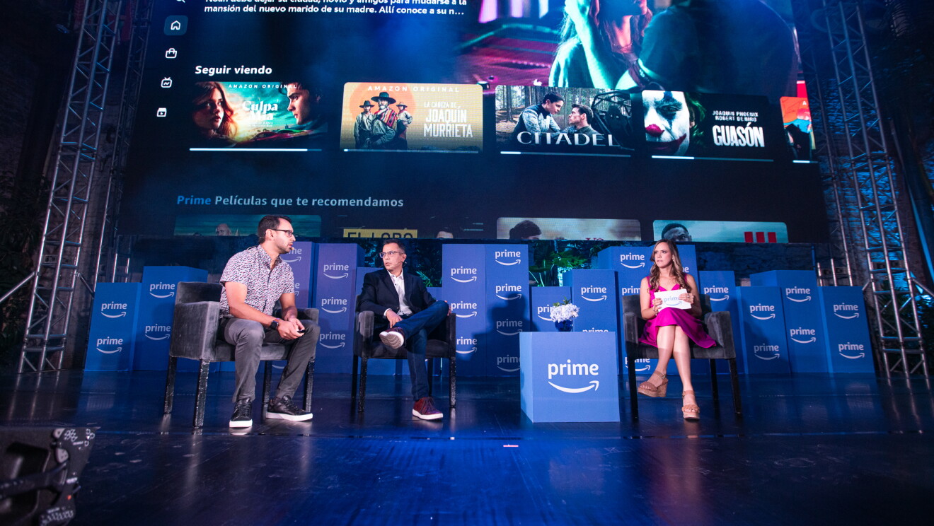 En un escenario con cajas con el logo de Prime, aparecn tres personas sentadas que no se pueden identificar y encima de ellas los carteles de las seires y películas de Prime Video en México como Culpa Mía, Citadel o La Cabeza de Joaquin Murrieta. 