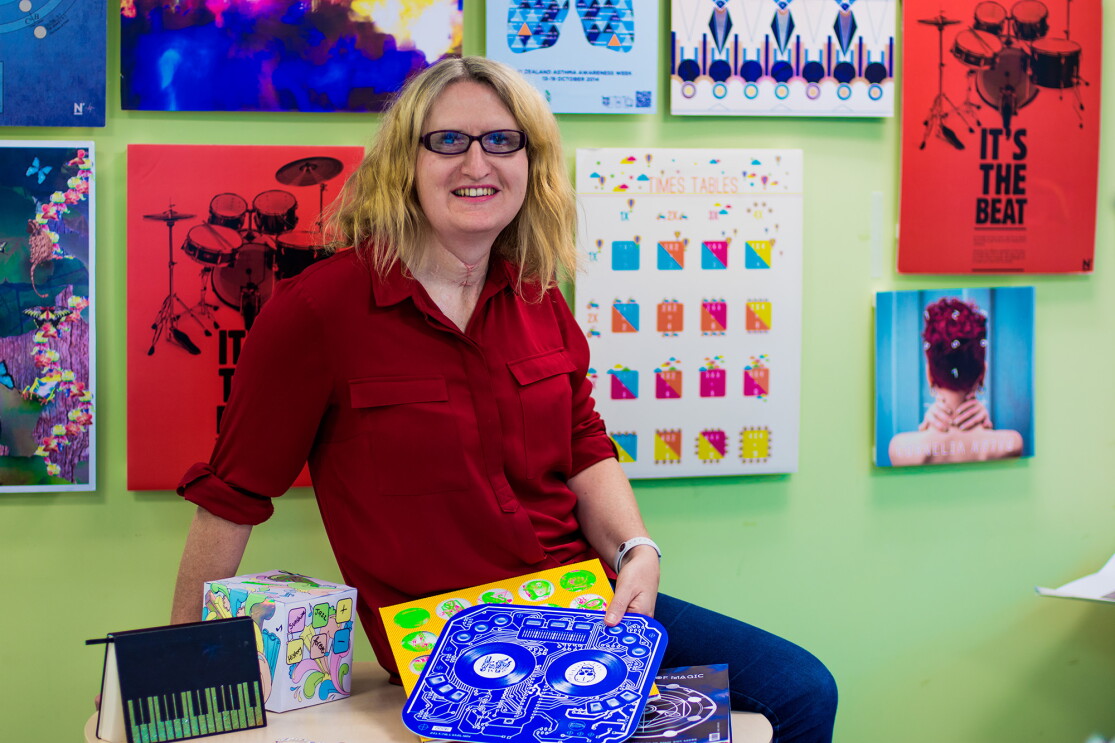 A blonde woman wearing glasses, jeans, and red button up shirt is seated on a tabletop in a room painted green, with myriad artwork hung on the wall.