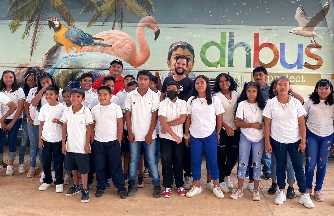 El DHBUS, un autobús acondicionado como aula itinerante, es un proyecto de innovación educativa diseñado para llevar programas a comunidades vulnerables y de difícil acceso. El bus está pintado con letras de colores dhbus project y pájaros exóticos típicos de México. Hay tres personas adultos que están en la parte trasera y delante, poco menos de 30 adolescentes vestidos con una camiseta blanca y jeans.