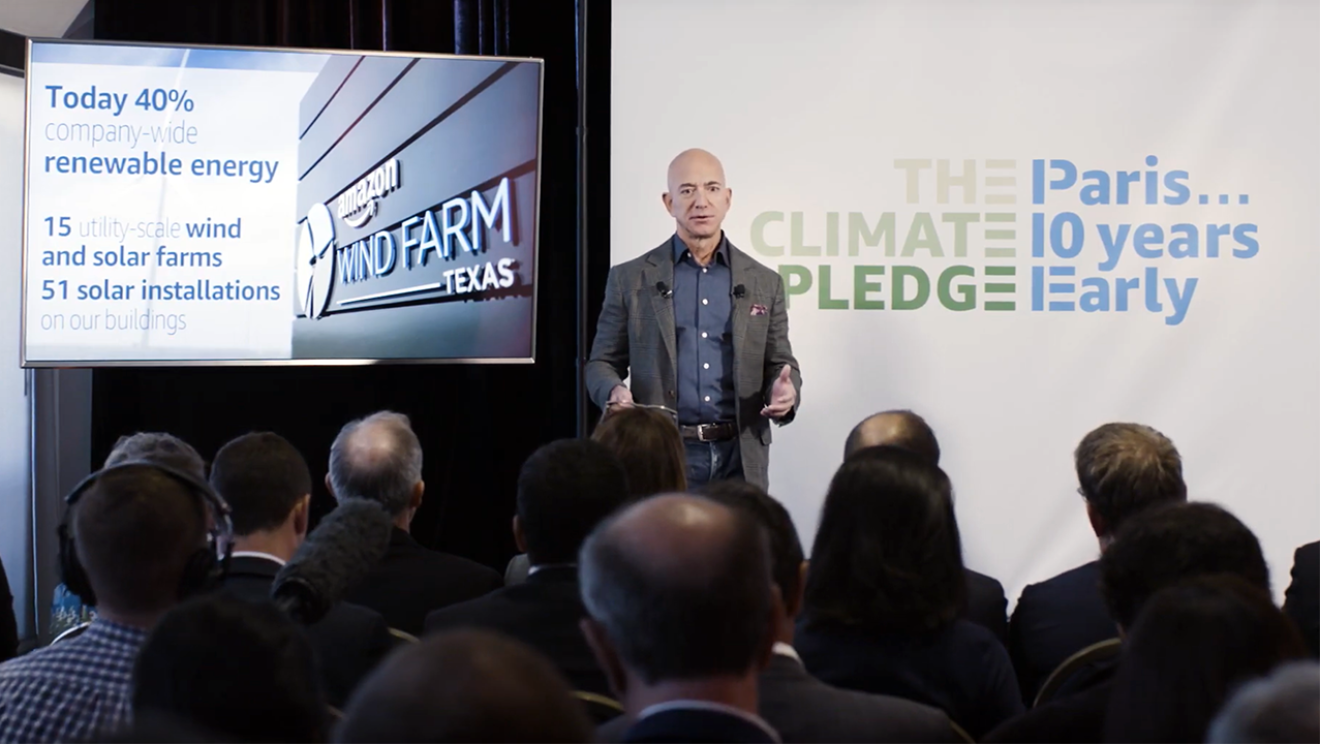Jeff Bezos stands at the front of a room, in front of a display that shares data points about renewable energy at Amazon. Also behind him is the logo for "The Climate Pledge."