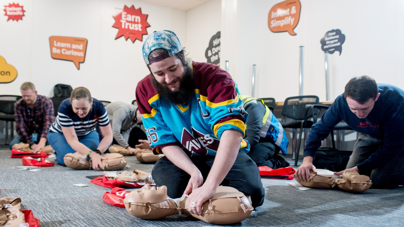 Amazon Operations Supervisor Lloyd Curley kneeling on the floor and practicing CPR on a dummy, along with five of his colleagues doing the same in the background.