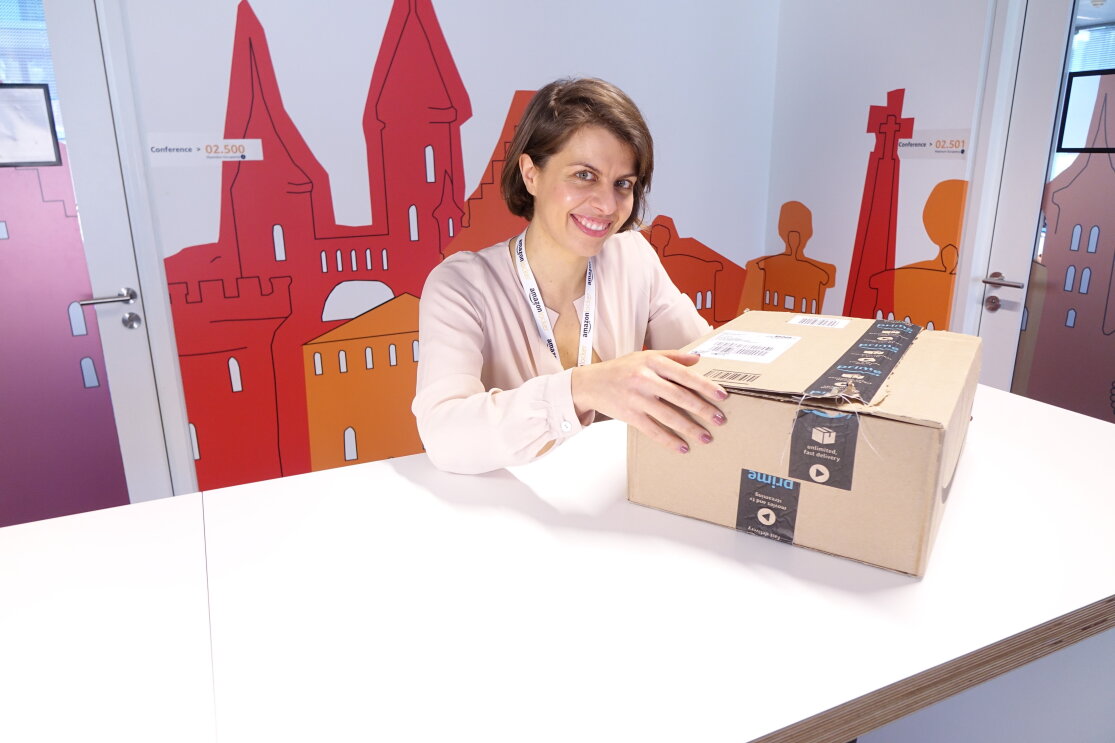 Ritratto di Marianna Pietrafesa, Responsabile europea del marketing per Amazon Locker e Pickup. La donna siede dietro un tavolo e poggia la mano destra su una scatola Amazon, poggiata di fronte a lei. Sullo sfondo un muro colorato nell'ufficio Amazon di Lussemburgo. 