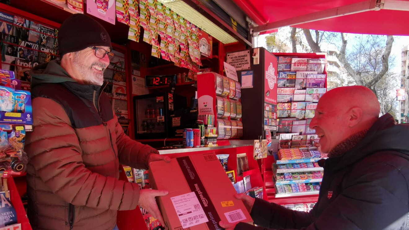 En un kiosco a pie de calle, en un día soleado, el quiosquero, un hombre alto de mediana edad que lleva barba y gafas y usa sombrero de lana, entrega un paquete a un cliente, un hombre también de mediana edad, que lleva un plumífero puesto. Hace sol, pero por el atuendo de las personas que aparecen en la foto, parece que es invierno. Ambos sonríen, parecen contentos. 