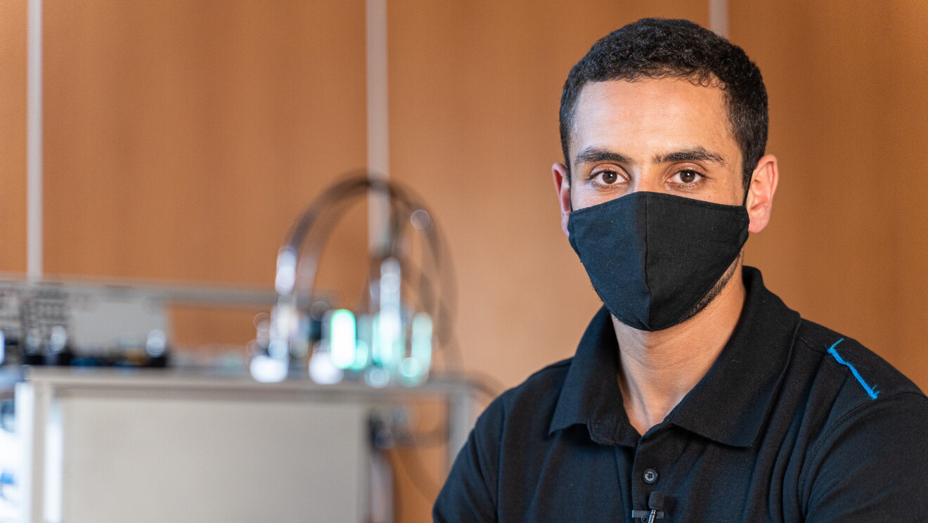 Nourddin El Bousouabi Llou, participante del programa de Aprendizaje de Amazon en España. Con un polo negro de Amazon, mira a cámara. Lleva mascarilla negra y tiene ojos grandes y marrones y el pelo corto y negro. De fondo se ve una mesa de trabajo con una máquina que no se puede apreciar. 