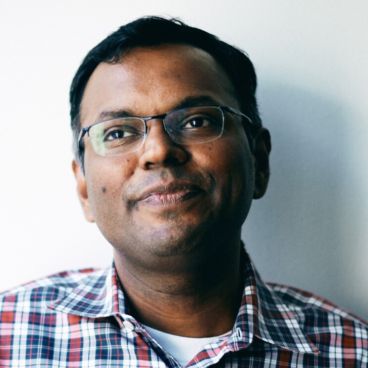 A close-cropped image of a man wearing a plaid button up over a white tee, and glasses sits in front of a white wall.