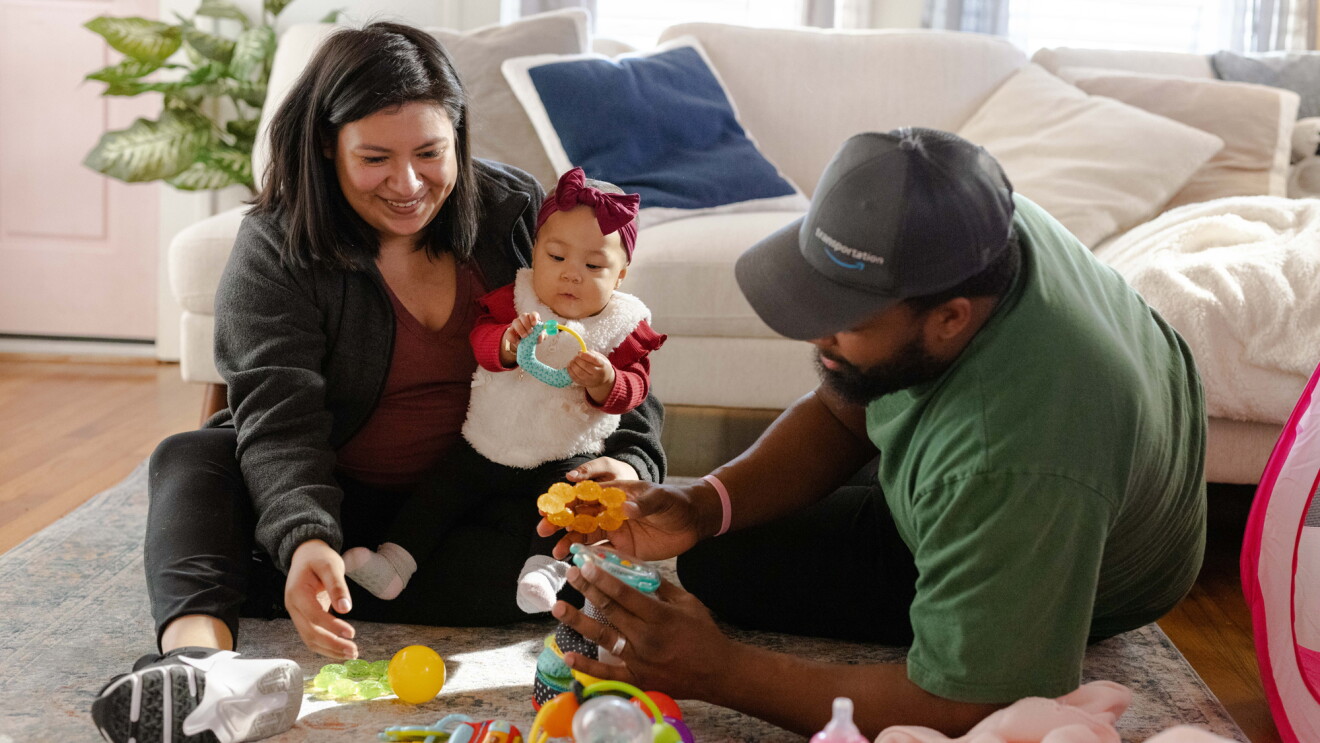 A woman sitting on a rug holds an infant next to a man holding baby toys.
