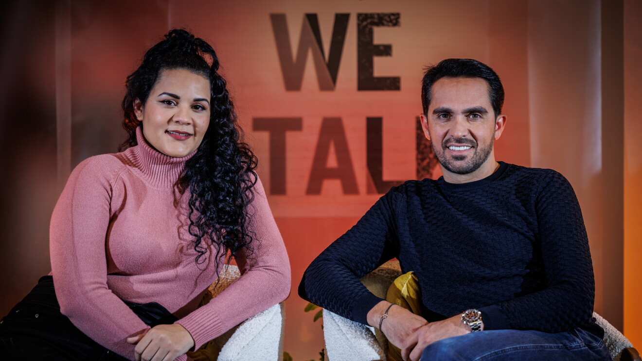 Leslie, trabajadora de Amazon, y Alberto Contador sentado en dos butacas blancas. Leslie va vestida con un jersey de color rosado de cuello alto y un pantalón negro de talle alto. Alberto Contador va con un jersey de punto azul y unos jeans de color azul también. Lleva un reloj en la mano izquierdo y una pulsera en la derecha. De fondo, en una pared de color naranja, está el lema de la campaña Yes, we talk. Leslie tiene el pelo largo negro rizado hasta el pecho. Los dos miran a la cámara sonriendo. 