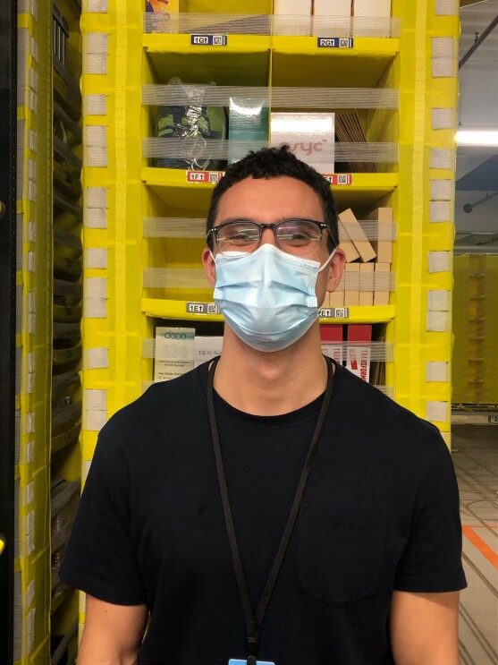 Lorcan Keating, an associate at Amazon's fulfilment centre in Durham, pictured at his work station with shelving pods in the background. He is smiling at the camera and wearing a face mask.