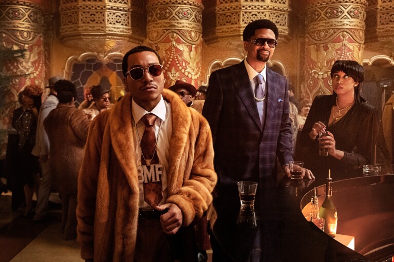 A still from Black Mafia Family, showing stylish men in luxurious attire at an opulent party with ornate golden pillars