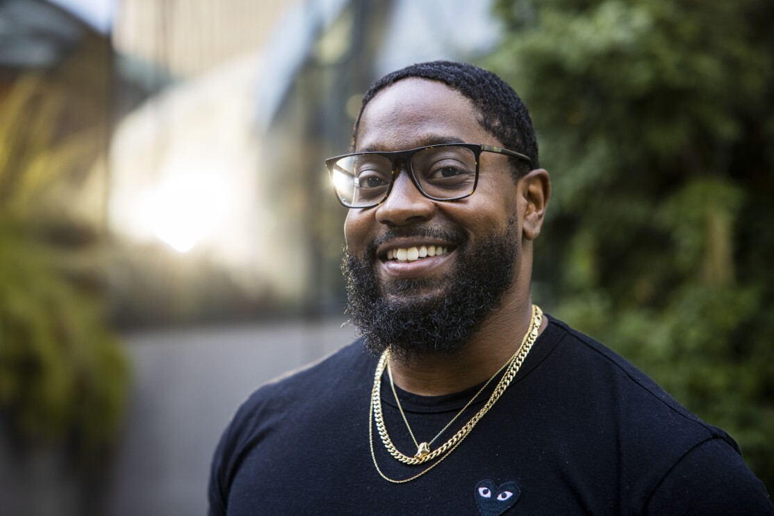 A smiling bearded man in a black T-shirt, glasses, and gold necklaces.