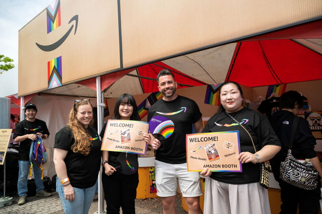 笑顔でブースの前に立つおそろいの黒いTシャツを着た4人の男女。そのうちの2人は「Welcome Amazon Booth」と書かれたプラカードを持っている