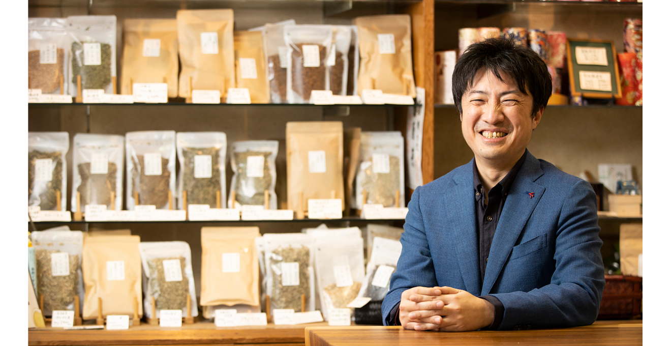 たくさんのお茶が並ぶ棚の前で、笑顔の川井さんの写真