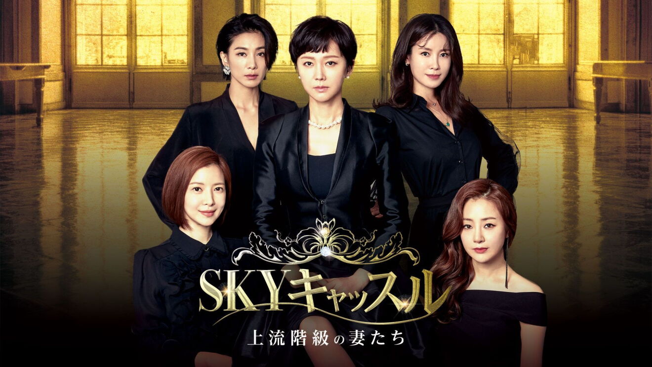 黒いフォーマルなスーツとドレスを着ている5人の女性が金色に輝く大広間でポーズをとっている