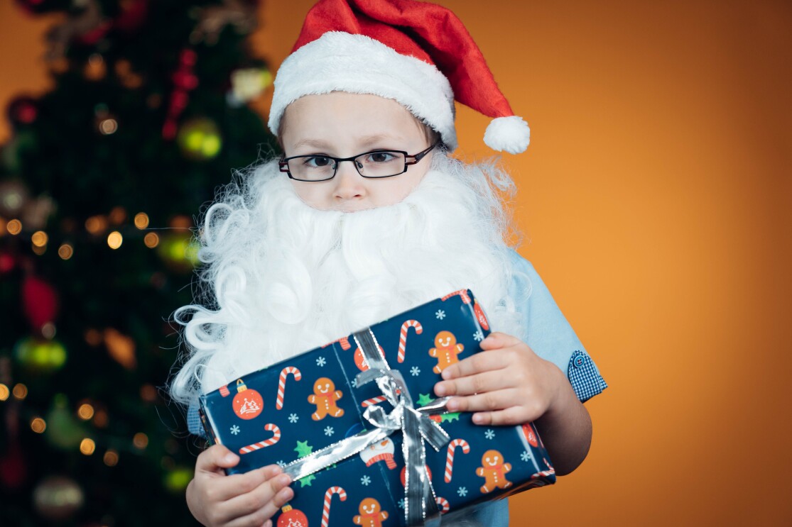 Ein Junge mit Brille, Weihnachtsmann-Mütze und weißem Weihnachtsmann-Bart schaut in die Kamera. Er hält ein blaues Geschenk in den Händen.