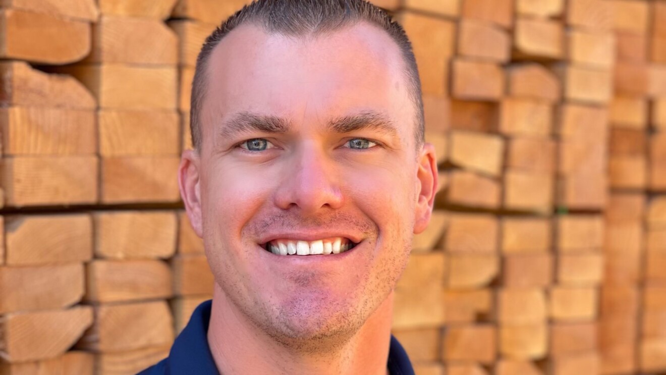 An image of a man smiling for a headshot photo. In the background there are stacks of wood pellets piled high.