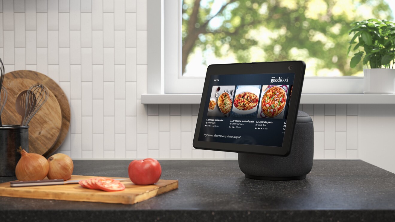 An image of the new Echo Show 10 on a kitchen counter. The device is showing recipes on BBC goodfood and next to the device there is a chopping board with tomatoes and onions.