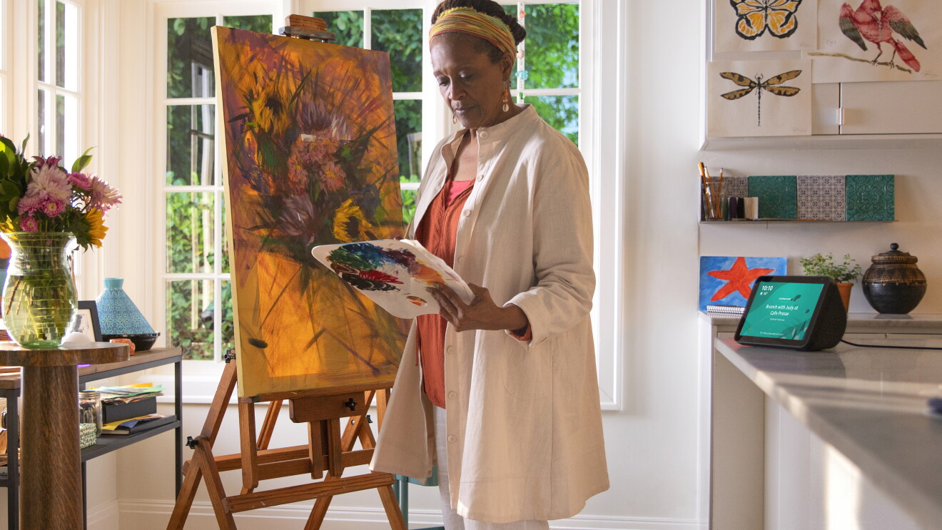 A mature woman works on a painting in her home. Behind her, an Alexa device is on the kitchen counter. 