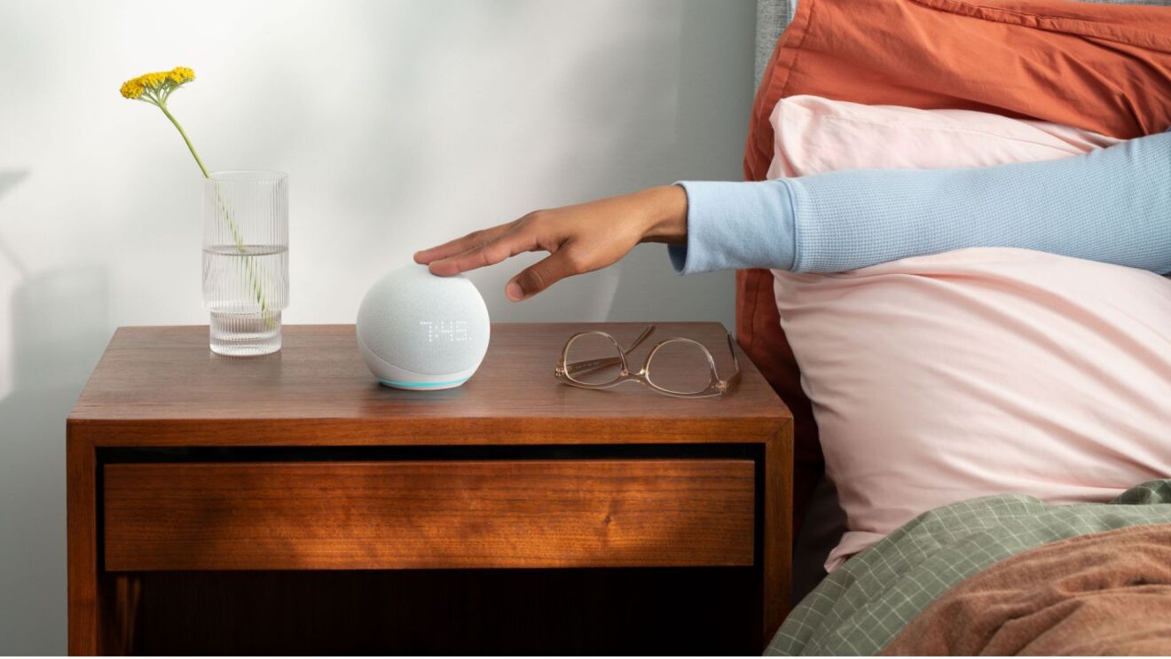Someone's hand reaching out Echo Dot on the bedside table from their bed.
