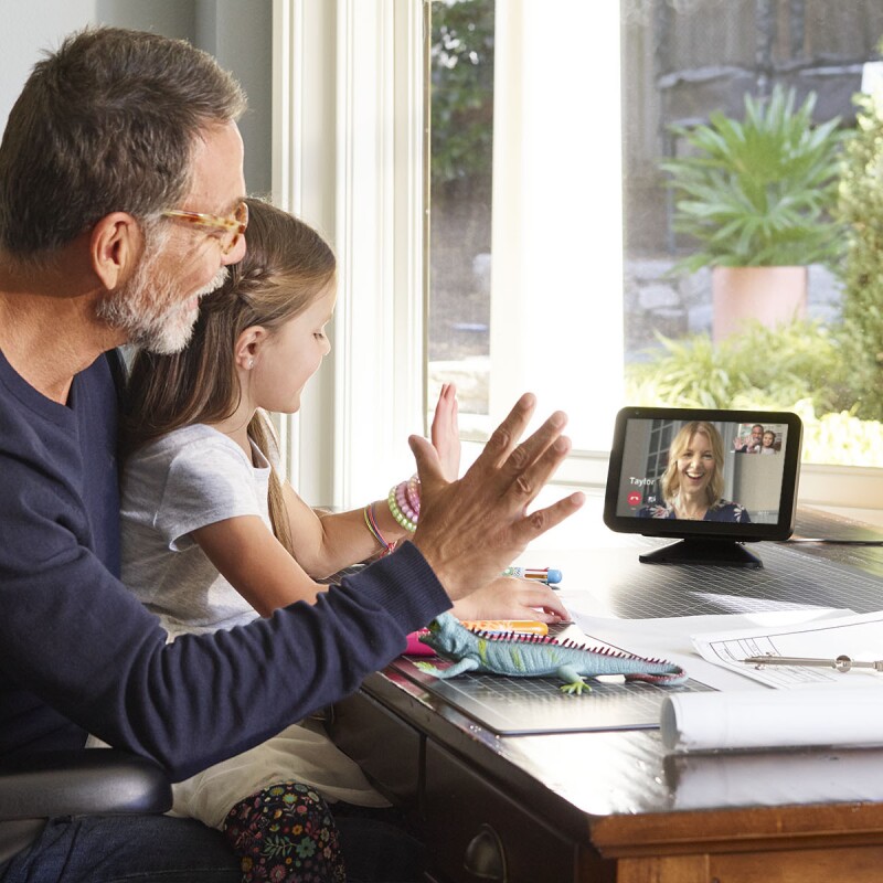 A little girl sits on a man's lap at a desk. They are both waving and looking at an Echo Show device, on which a woman is smiling and engaging with them.