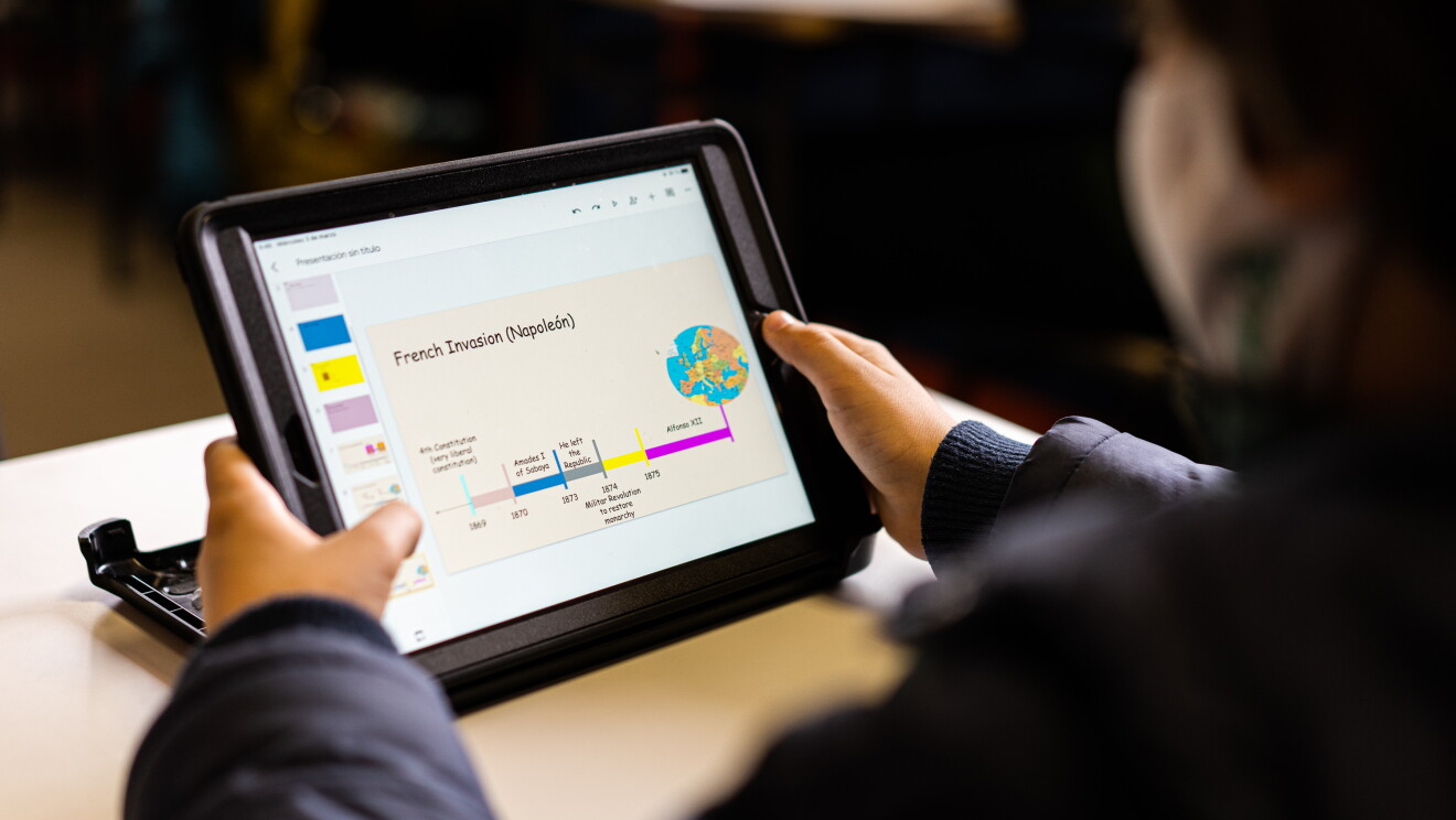 Amazon Digital Lab es un mundo de recursos digitales gratuitos para usar en el aula y en casa. Un plano desenfocado de un alumno con una tableta en la mano y habla de historia, de la invasión de Napoleón, en inglés.  El chico agarra la tableta con las manos. 