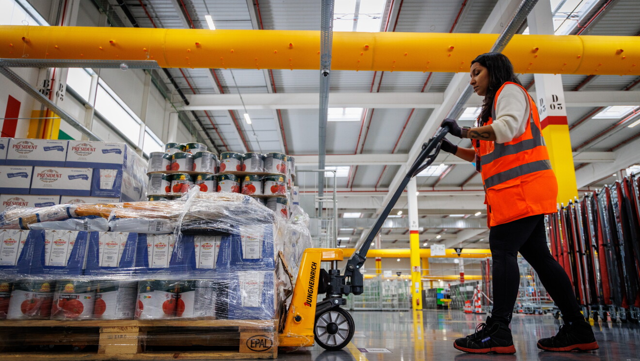 Dentro de un centro logístico de Amazon aparece una mujer morena, con el pelo largo, pantalón negro, jersey blanco, zapatos con proyección y un chaleco naranja con un toro semiautomático. En el palé se ven productos que no se pueden identificar y que están envueltos de papel transparente. 