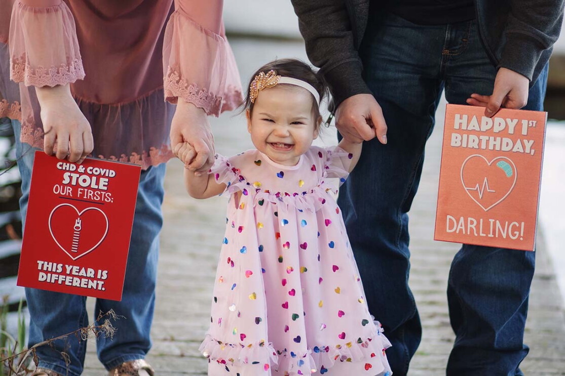 A toddler stands holding her mom and dad's hands. Her parents hold large greeting cards on either side of her. 